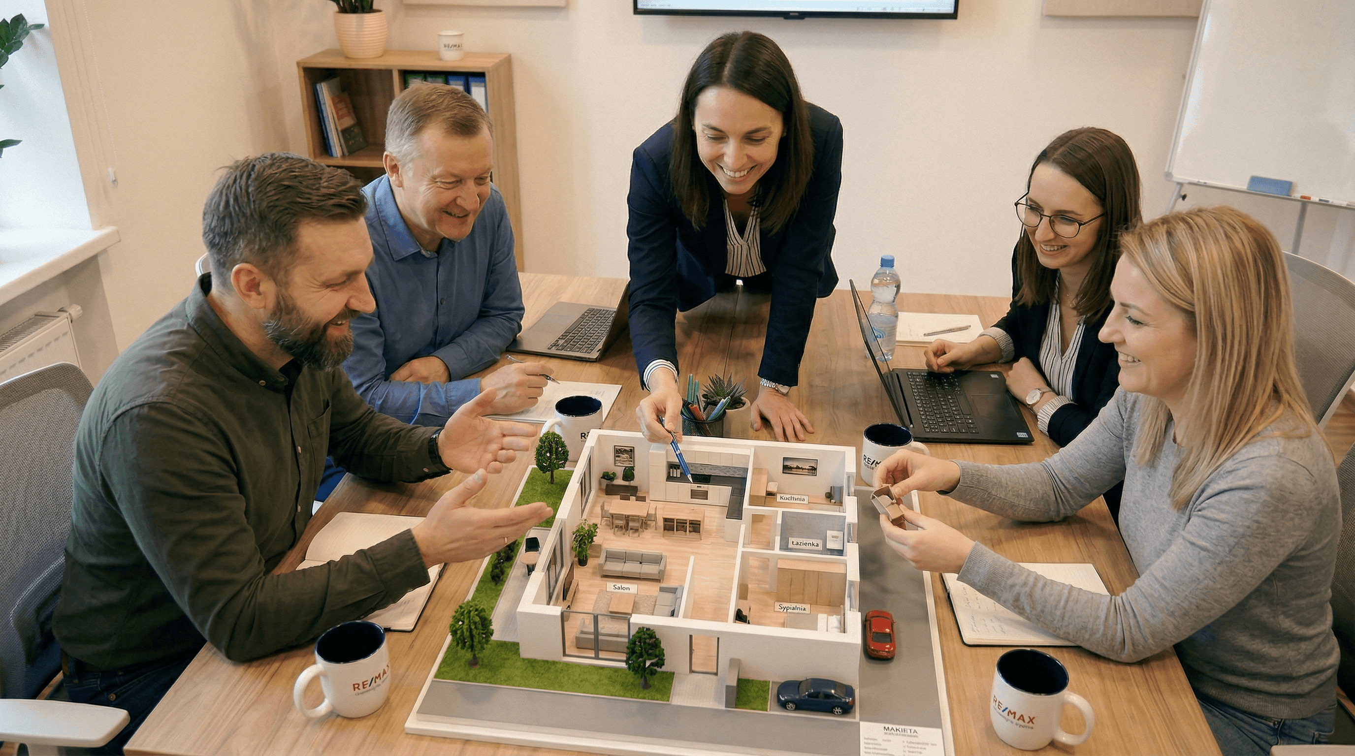 Zespół REMAX Unity omawia ofertę przy stole - pięć osób analizuje makietę mieszkania i rozmawia nad detalami.